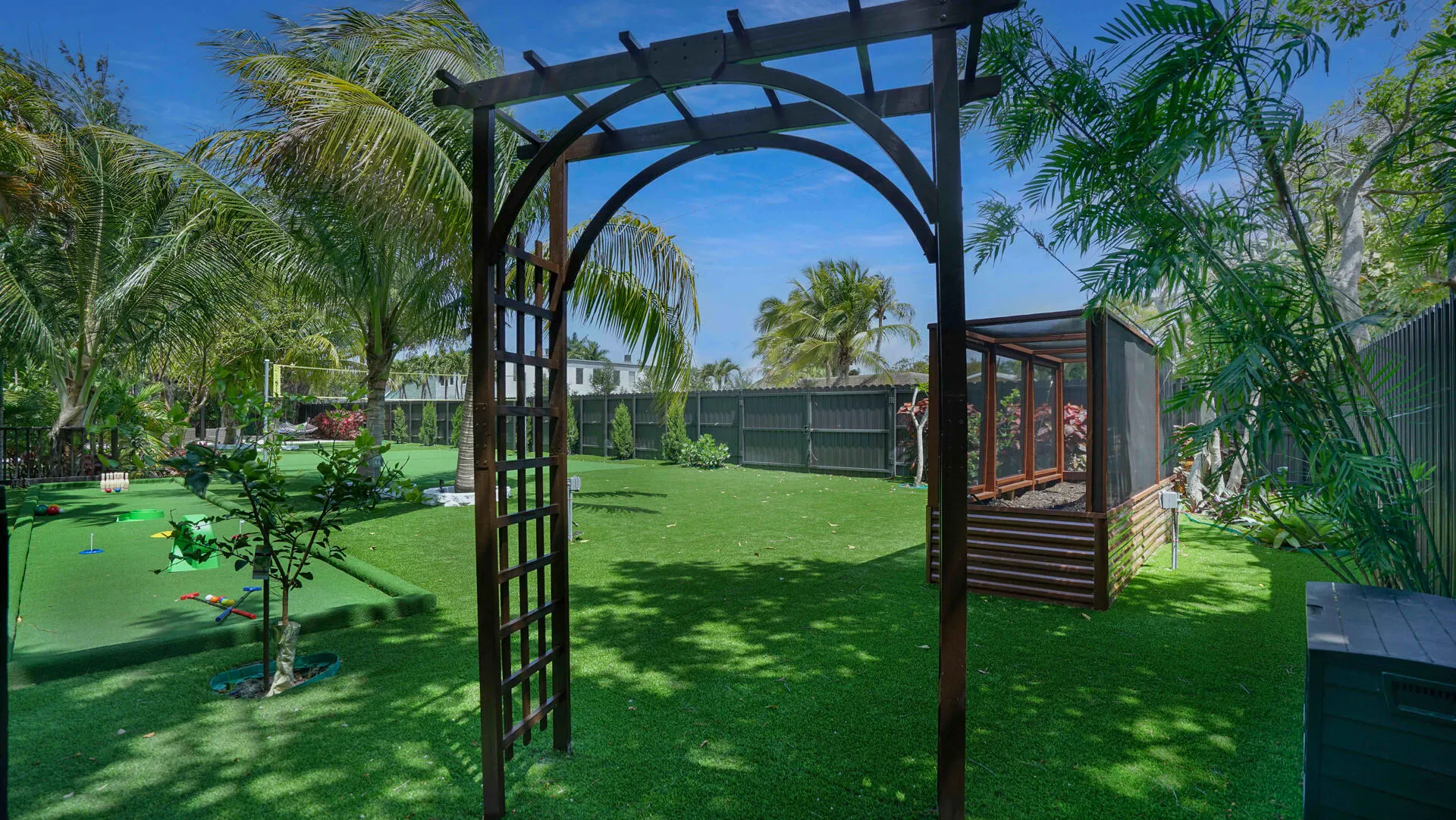 Expansive backyard featuring a central wooden arbor, lush artificial turf, a putting green, mature palm trees, and a raised garden bed with a screened enclosure