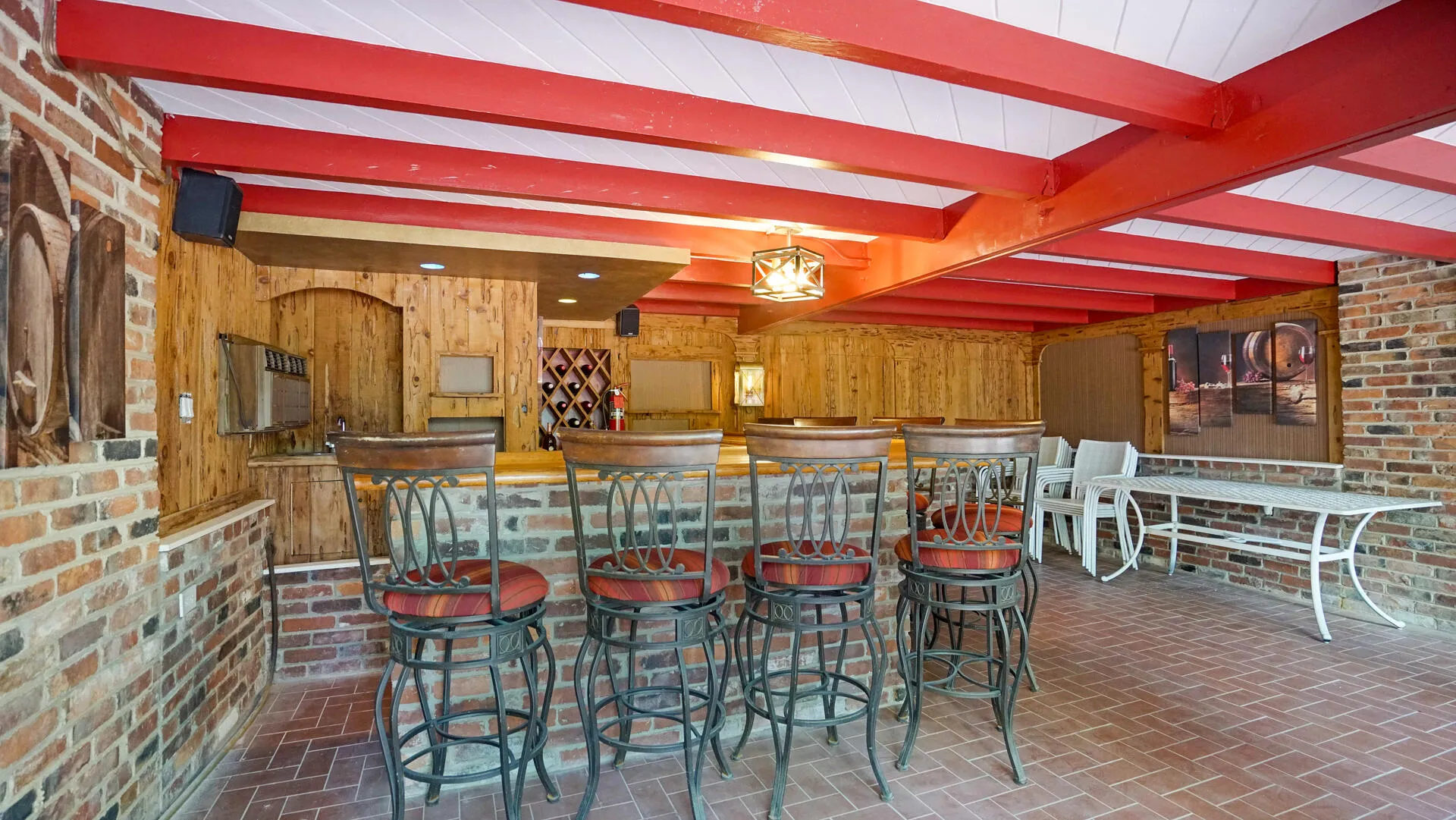Indoor-outdoor entertainment area featuring brick-patterned flooring, exposed red ceiling beams, and a built-in bar with wood-finish paneling