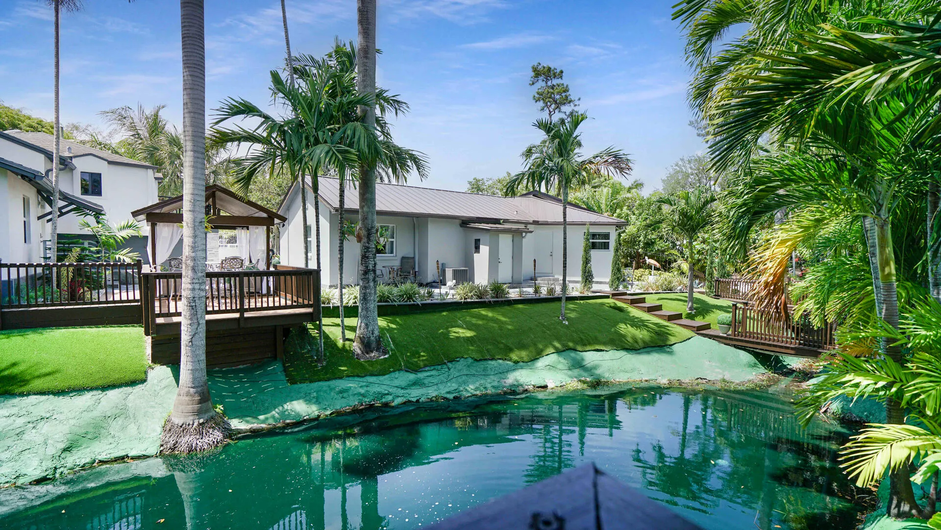 Waterside property with mature palm trees, featuring a wooden deck with a built-in gazebo, and a secondary structure with a metal roof