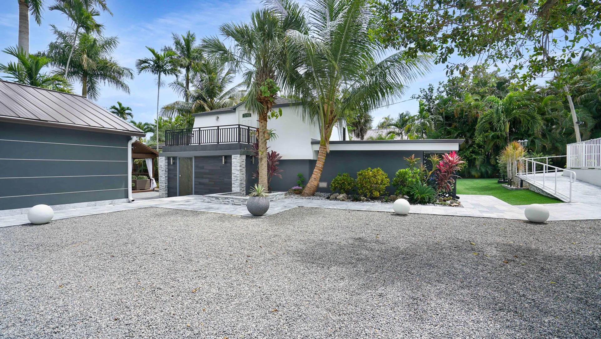 Modern exterior featuring a standing seam metal roof, stone accents, a second-story balcony with metal railings, a paved walkway, and mature palm trees