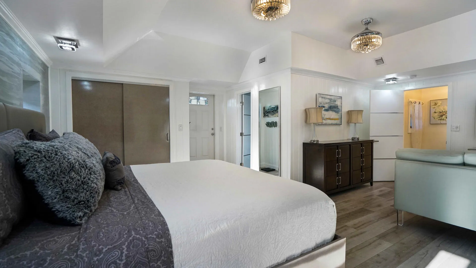 Expansive room featuring wood-finish flooring, two ceiling-mounted chandelier light fixtures, white paneling, and an entry door with glass inserts