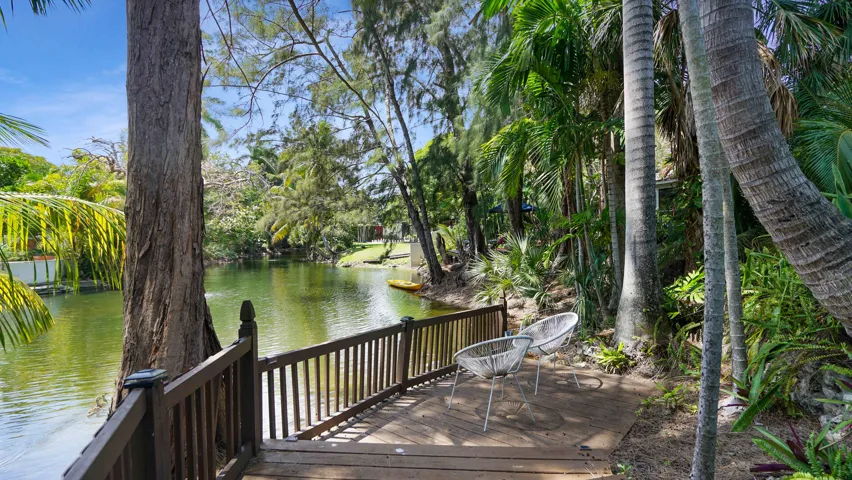 Waterfront wood-finish deck featuring a slatted railing, surrounded by lush tropical foliage and mature trees, with direct water access
