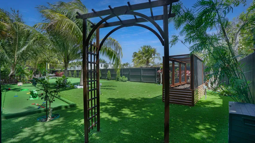 Expansive backyard featuring a central wooden arbor, lush artificial turf, a putting green, mature palm trees, and a raised garden bed with a screened enclosure
