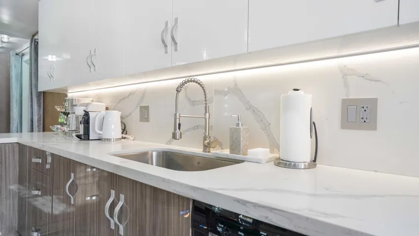 Kitchen featuring a large stainless steel sink with a pull-down faucet, under-cabinet lighting, white upper cabinetry with curved handles, wood-finish lower cabinetry, and a light-toned countertop with a matching backsplash