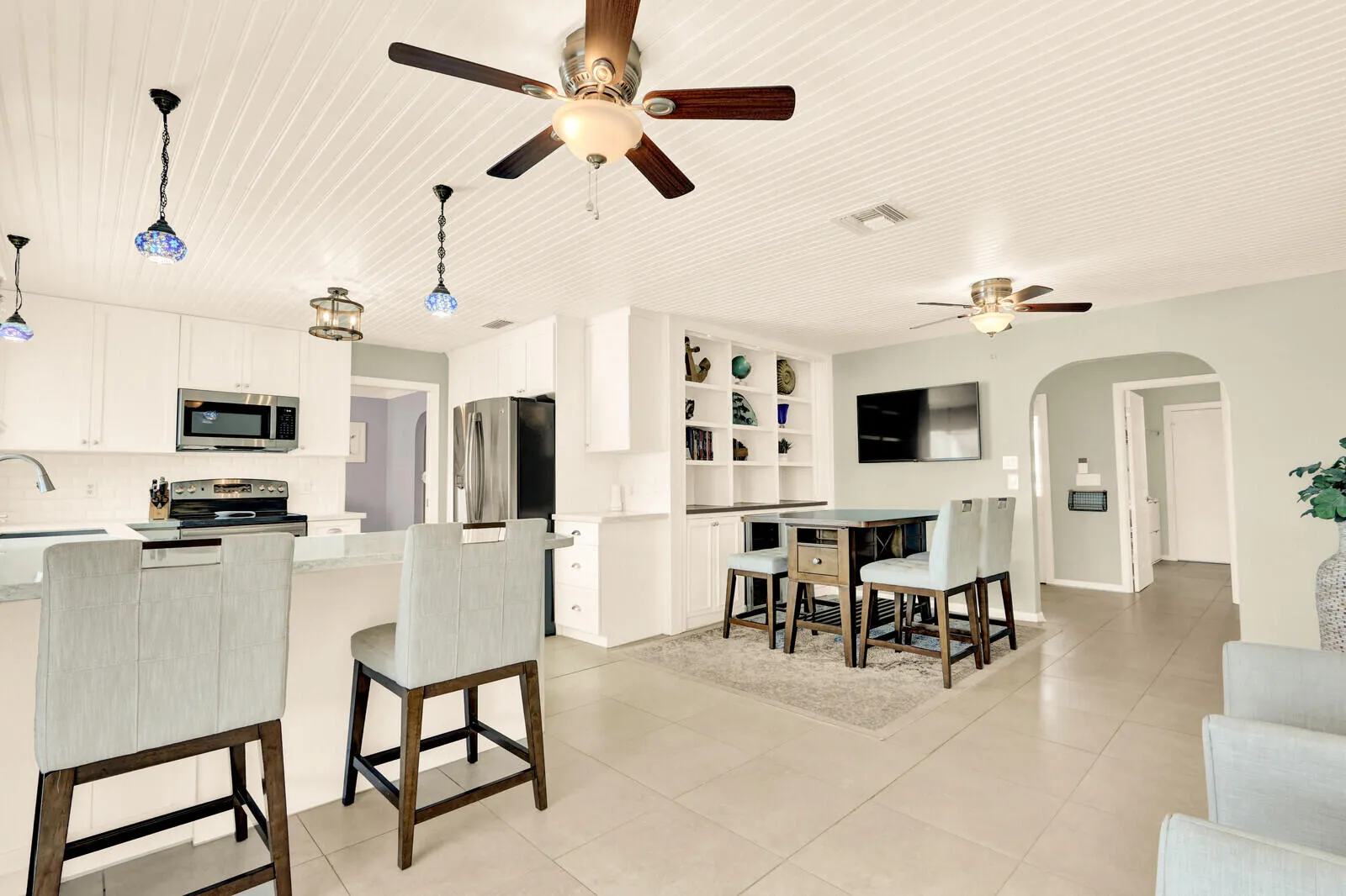 Open-concept kitchen and dining area featuring white shaker cabinetry, stainless steel appliances, a breakfast bar with stone-finish countertops, tile flooring, and a shiplap ceiling with multiple ceiling fans