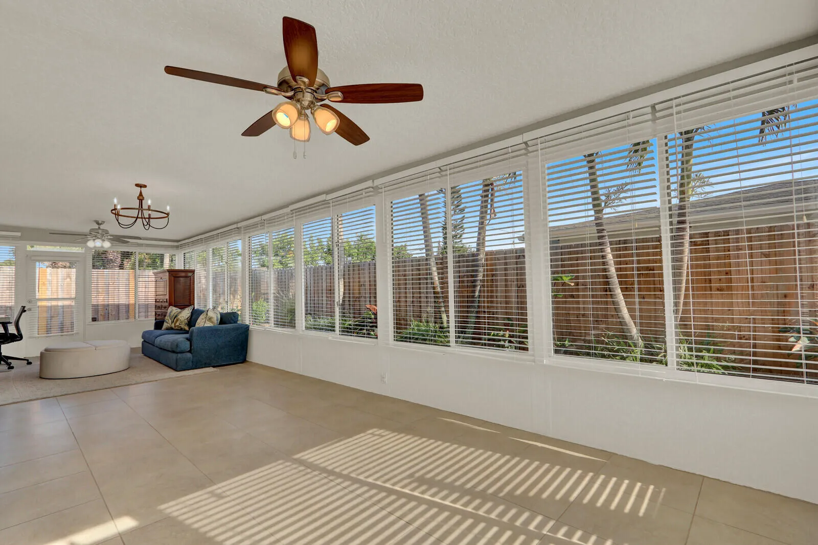 Spacious room featuring light-toned tile flooring, multiple large windows with horizontal blinds, and a ceiling fan with integrated lighting