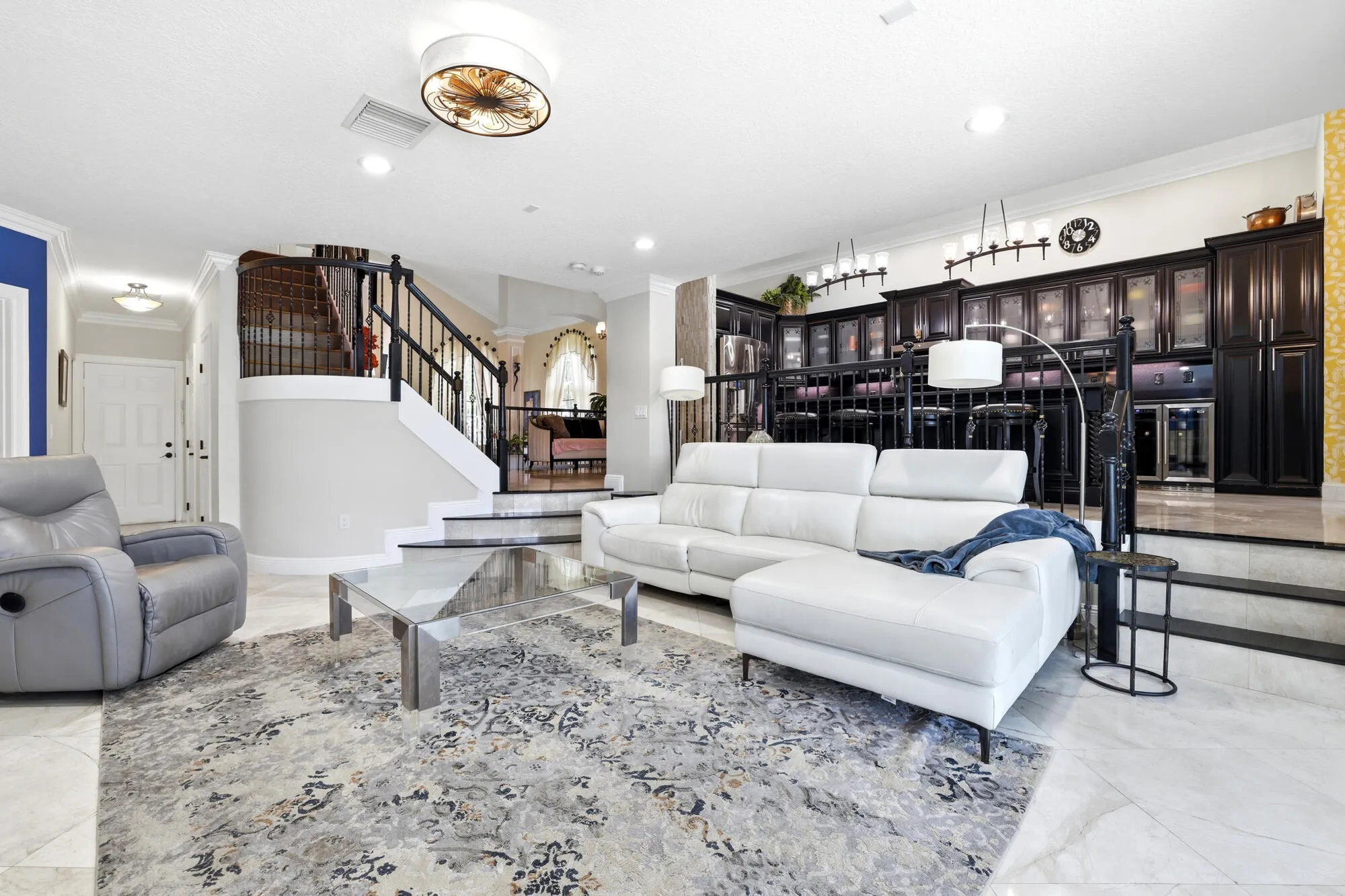 Spacious living area featuring light-toned tile flooring, a built-in wet bar with dark cabinetry, and an elegant staircase with dark wood-finish handrails