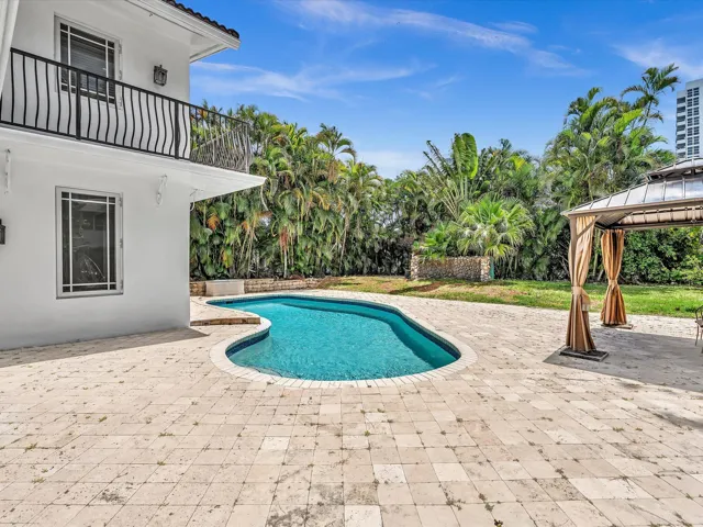 Resort-style backyard featuring a freeform swimming pool with stone coping, an expansive paver patio, and mature tropical landscaping
