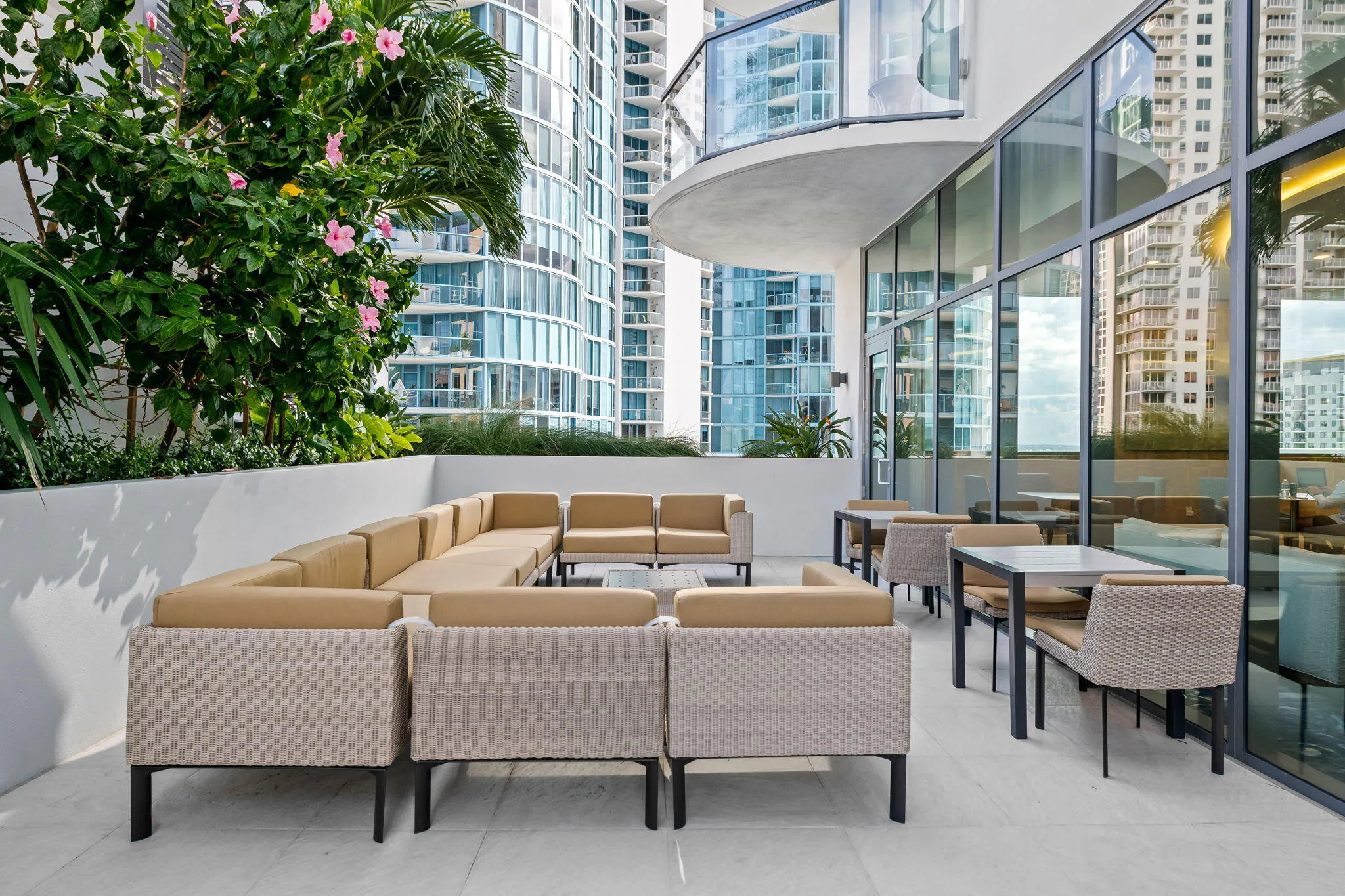 Spacious outdoor terrace featuring a large sectional seating area, additional dining tables, floor-to-ceiling glass paneling, and integrated planters with established foliage