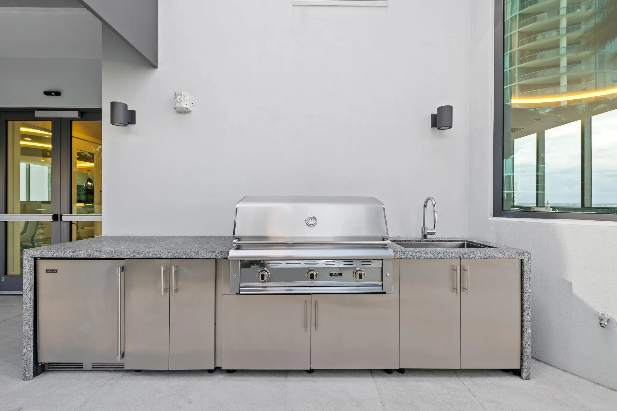 Outdoor kitchen featuring an integrated stainless steel grill, a stainless steel under-counter refrigerator, a stainless steel sink with a gooseneck faucet, and a stone-finish countertop