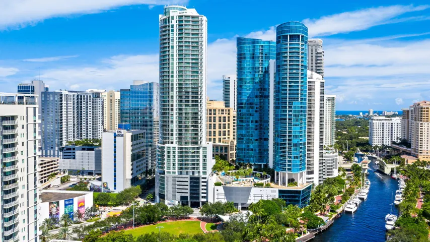 Skyline featuring multiple high-rise structures, a waterway with docked boats, abundant green spaces, and distant ocean views