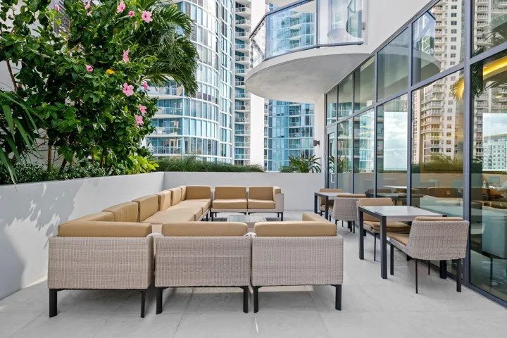Spacious outdoor terrace featuring a large sectional seating area, additional dining tables, floor-to-ceiling glass paneling, and integrated planters with established foliage