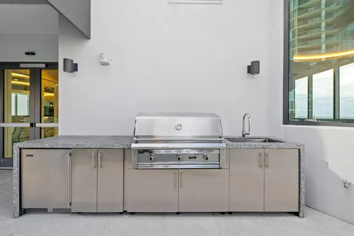 Outdoor kitchen featuring an integrated stainless steel grill, a stainless steel under-counter refrigerator, a stainless steel sink with a gooseneck faucet, and a stone-finish countertop