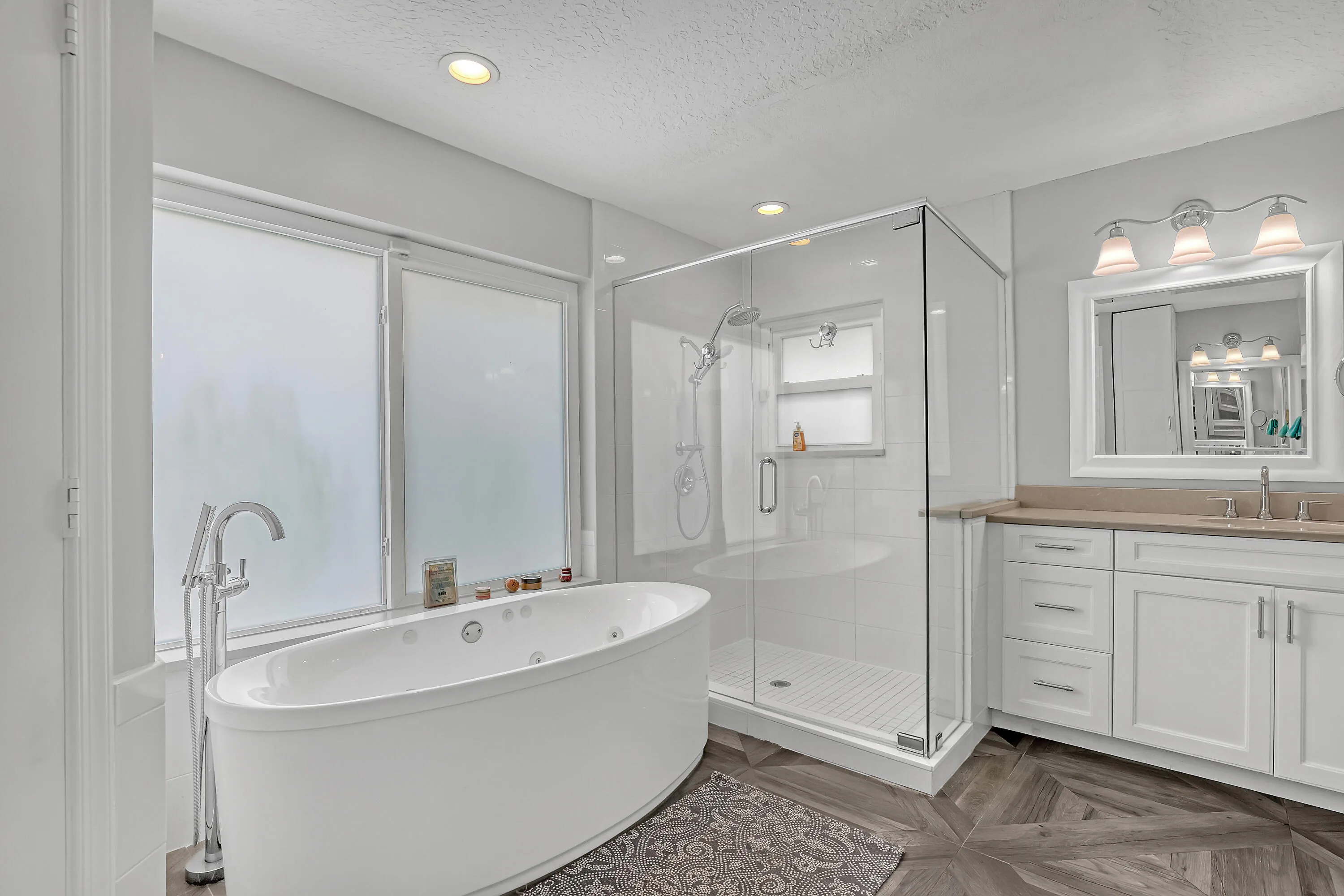 Contemporary bathroom featuring a freestanding soaking tub with floor-mounted faucet, a glass-enclosed shower with tiled walls, and a double vanity with white cabinetry and light-toned countertops
