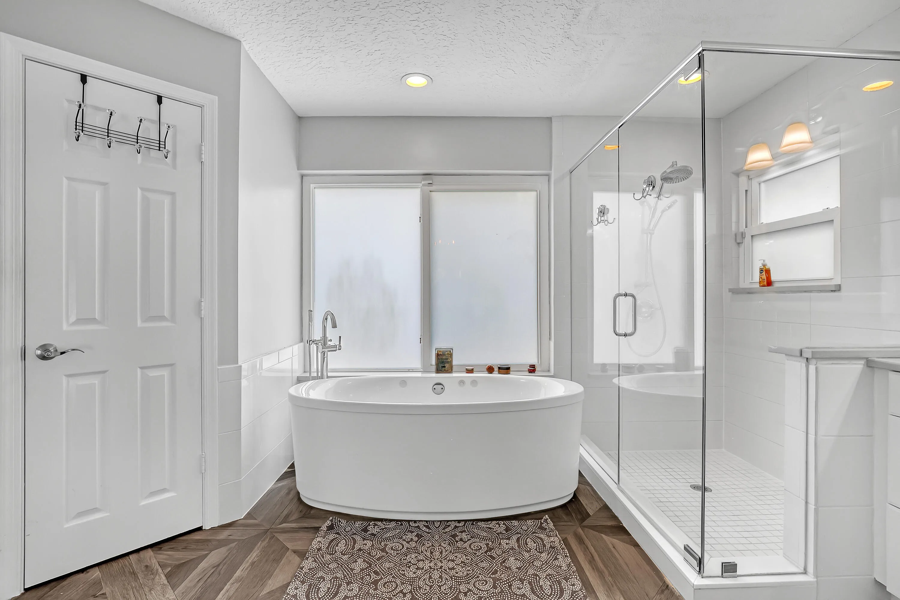 Freestanding oval soaking tub with floor-mounted faucet, glass-enclosed shower with subway tile surround, frosted windows, and wood-finish flooring
