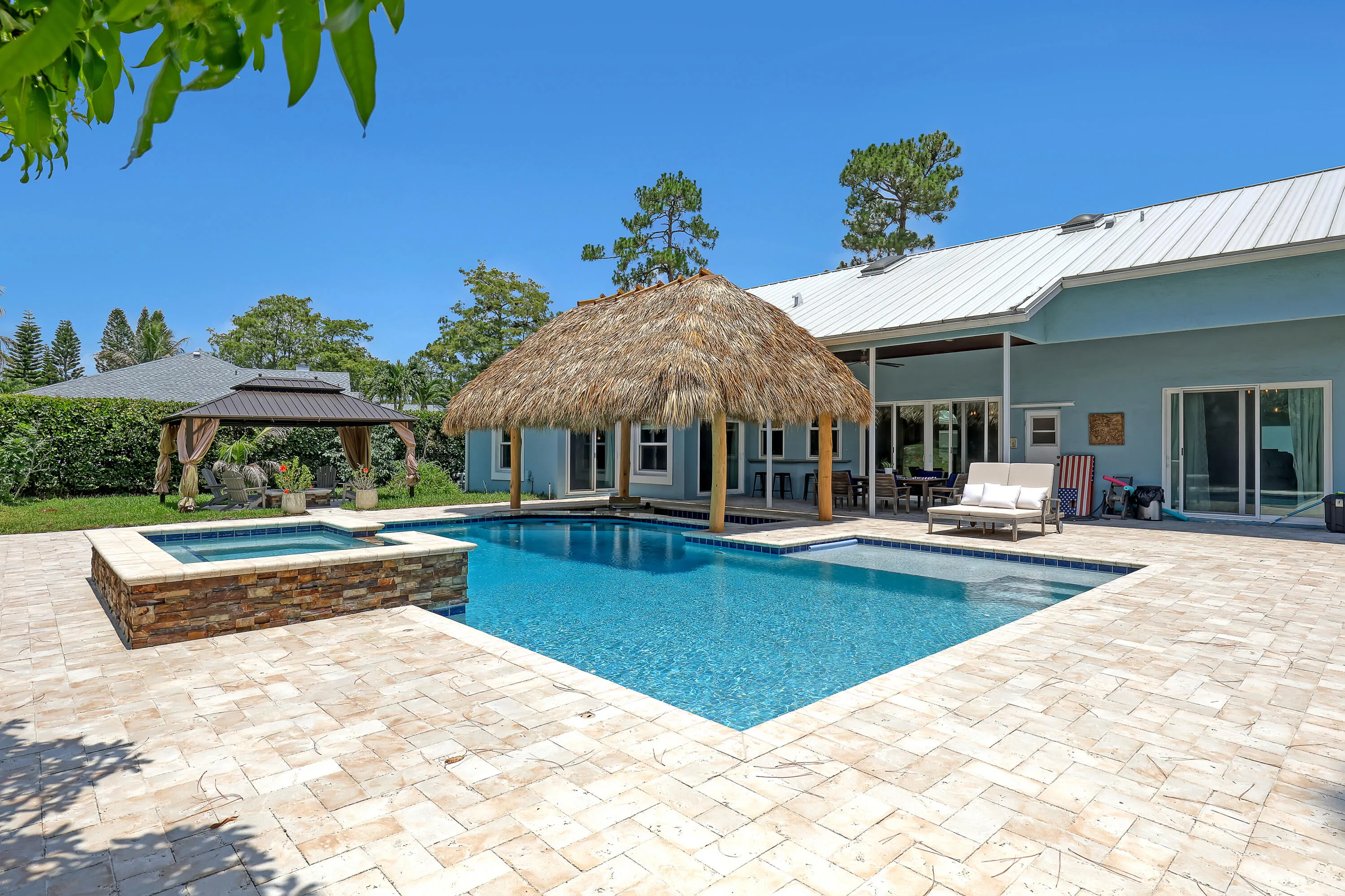 Expansive paver patio with a rectangular pool, integrated spa with stone surround, and a thatched-roof cabana