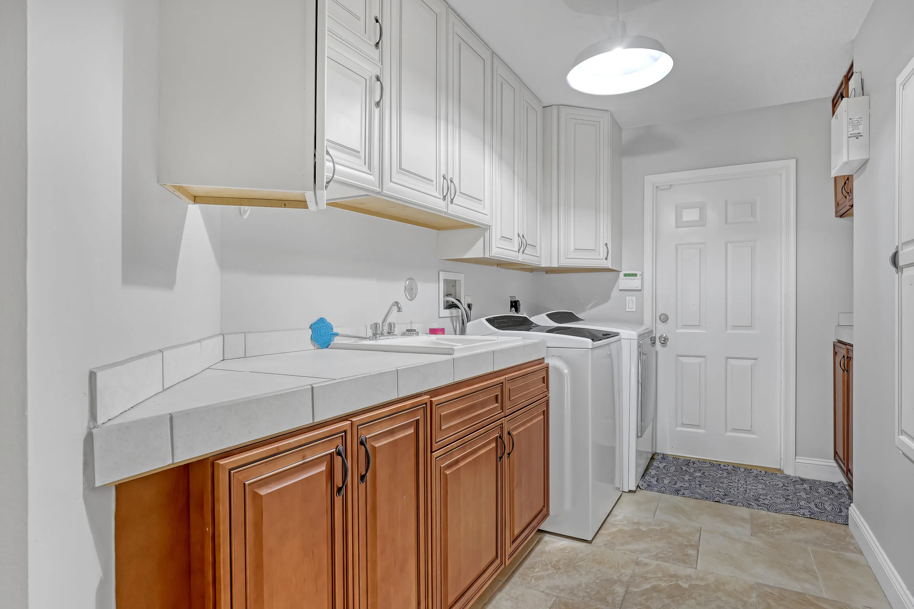 Utility area featuring white upper cabinetry, wood-finish lower cabinetry, a tiled countertop with an integrated sink, and durable tile flooring