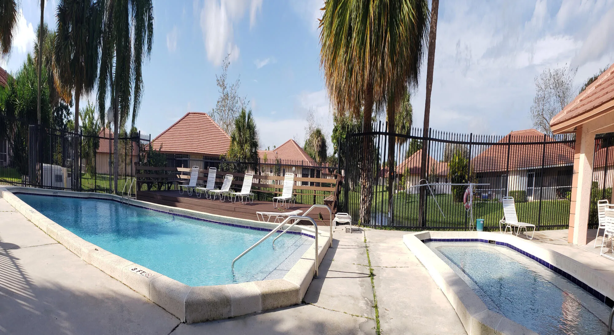 Community swimming pool and separate spa featuring a surrounding concrete deck, black metal fencing, and mature palm trees