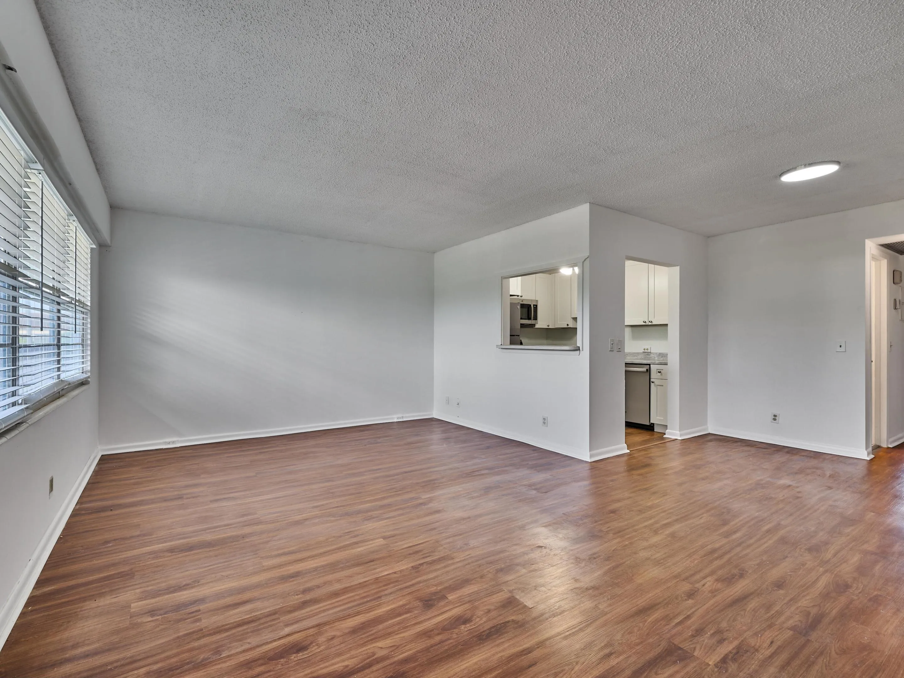Open concept living area featuring wood-finish flooring, white walls, and a large window with horizontal blinds