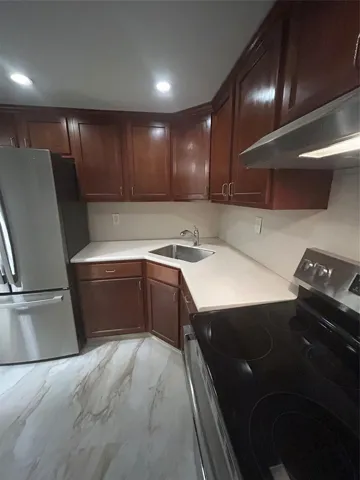 recessed lighting from a dimmer switch shine on wood cabinets above and below. New appliances include refrigerator, stove and hood. White marble counter tops with steel sink and hosed faucet have lights under cabinets. Steel appliances on ceramic tile with similar vein colored flooring.