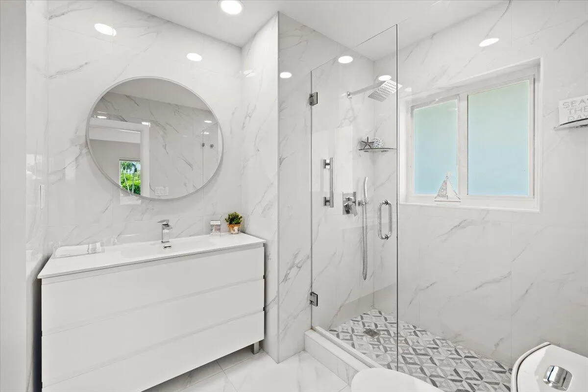 Bathroom featuring a glass-enclosed shower, rain showerhead, patterned shower floor tile, white vanity with integrated sink, and a circular mirror