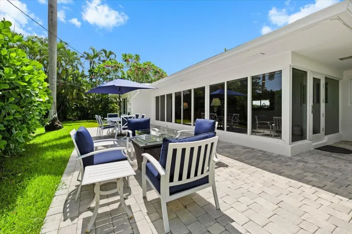 Expansive paver patio with a manicured lawn, lush tropical landscaping, floor-to-ceiling windows, and a glass-paneled exterior door