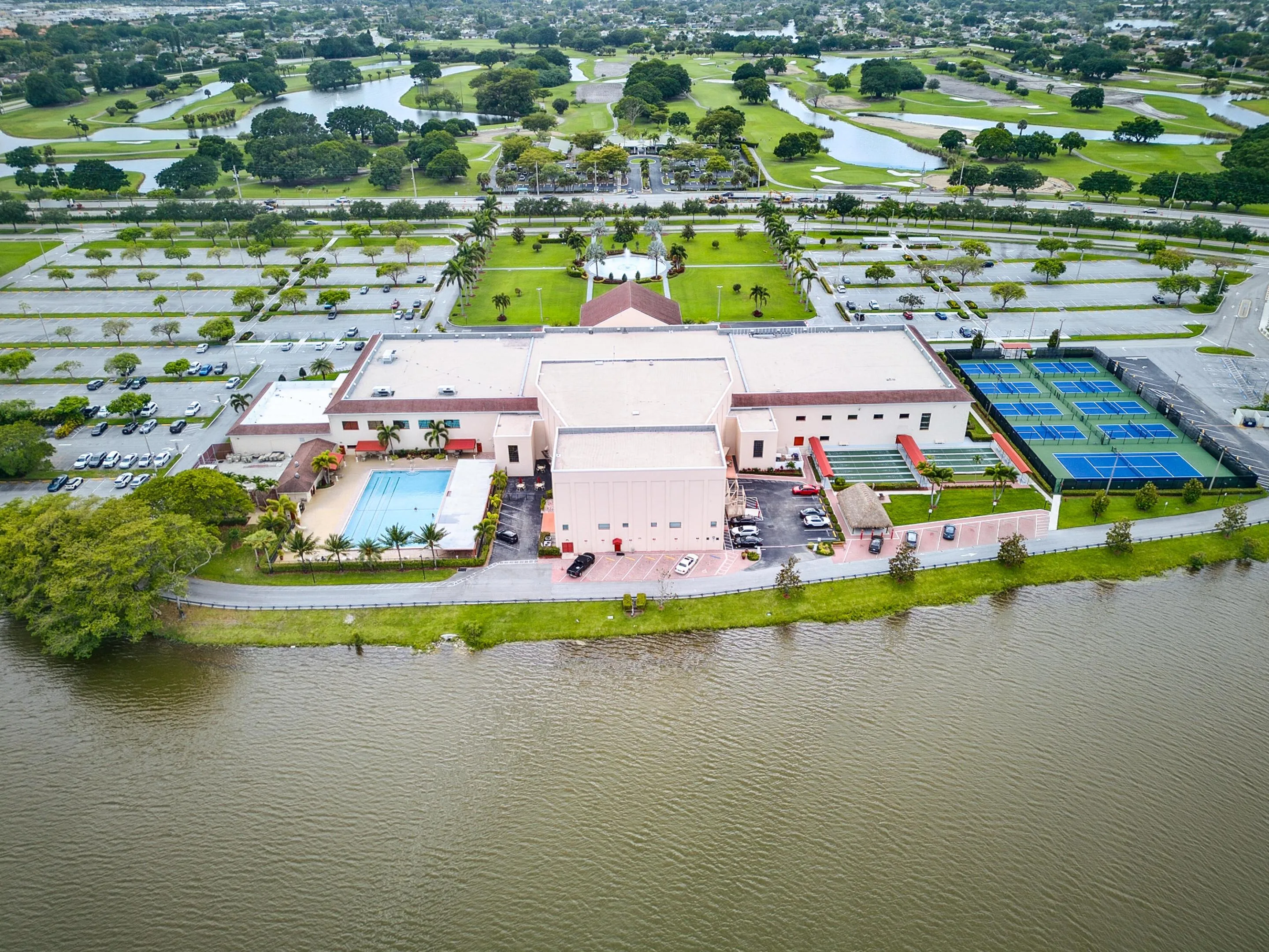 Expansive property featuring a large main building, multiple tennis courts, an outdoor swimming pool, and extensive parking facilities, bordered by a substantial body of water