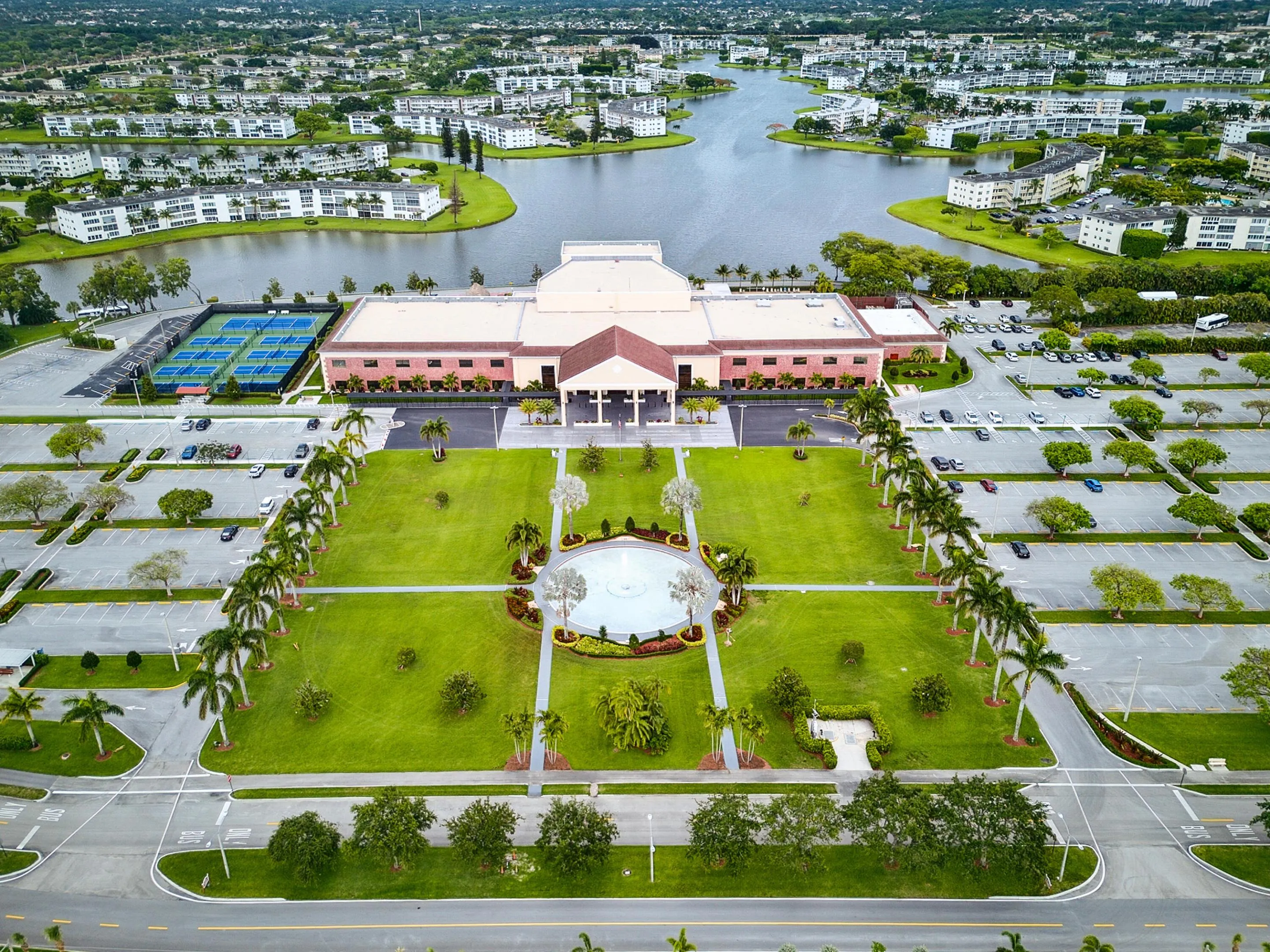 Expansive community grounds featuring a central building with a covered entrance, a water feature, multiple tennis courts, and ample parking