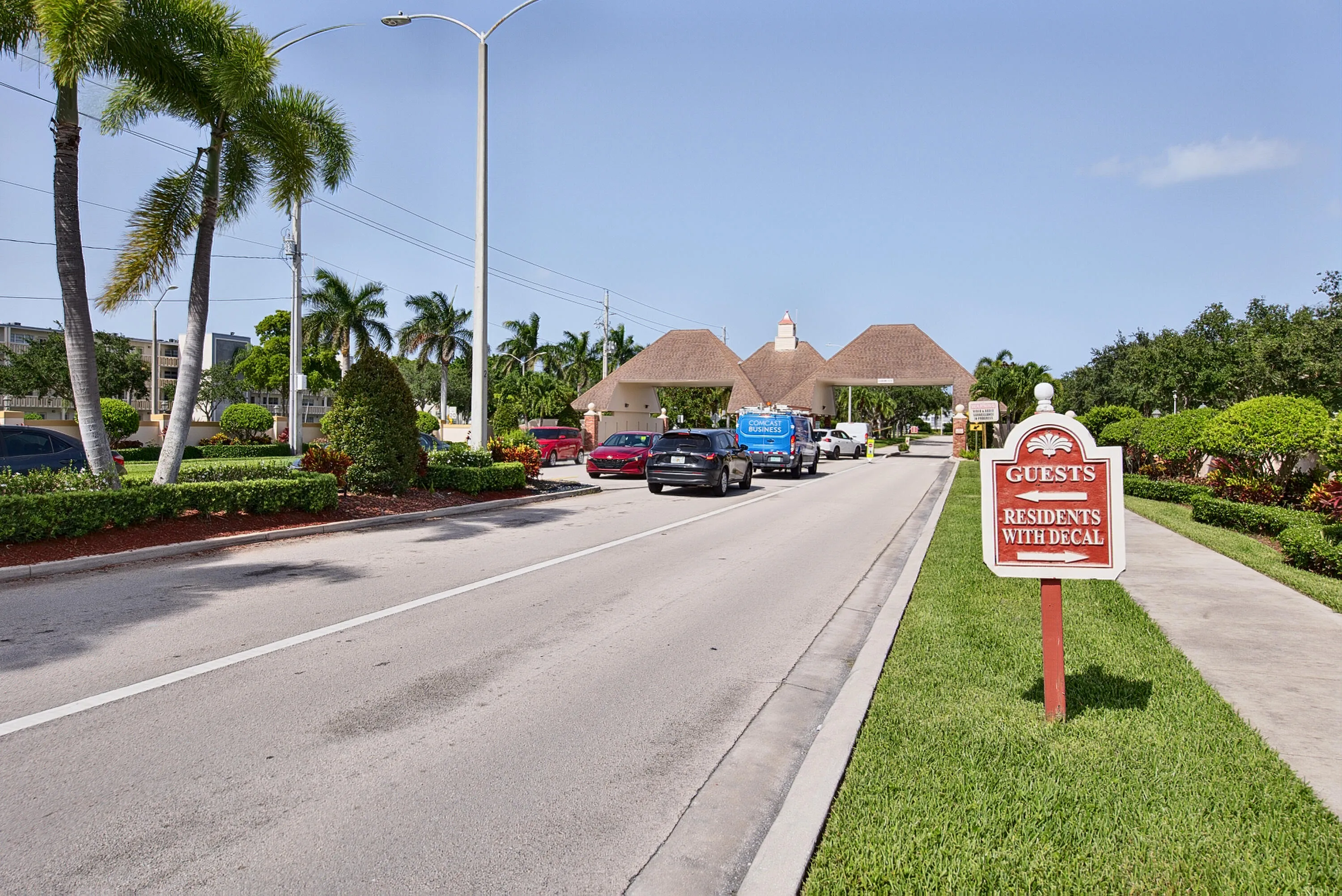 Gated entry featuring a multi-lane access point, thatched roof architectural elements, extensive perimeter landscaping, and mature palm trees