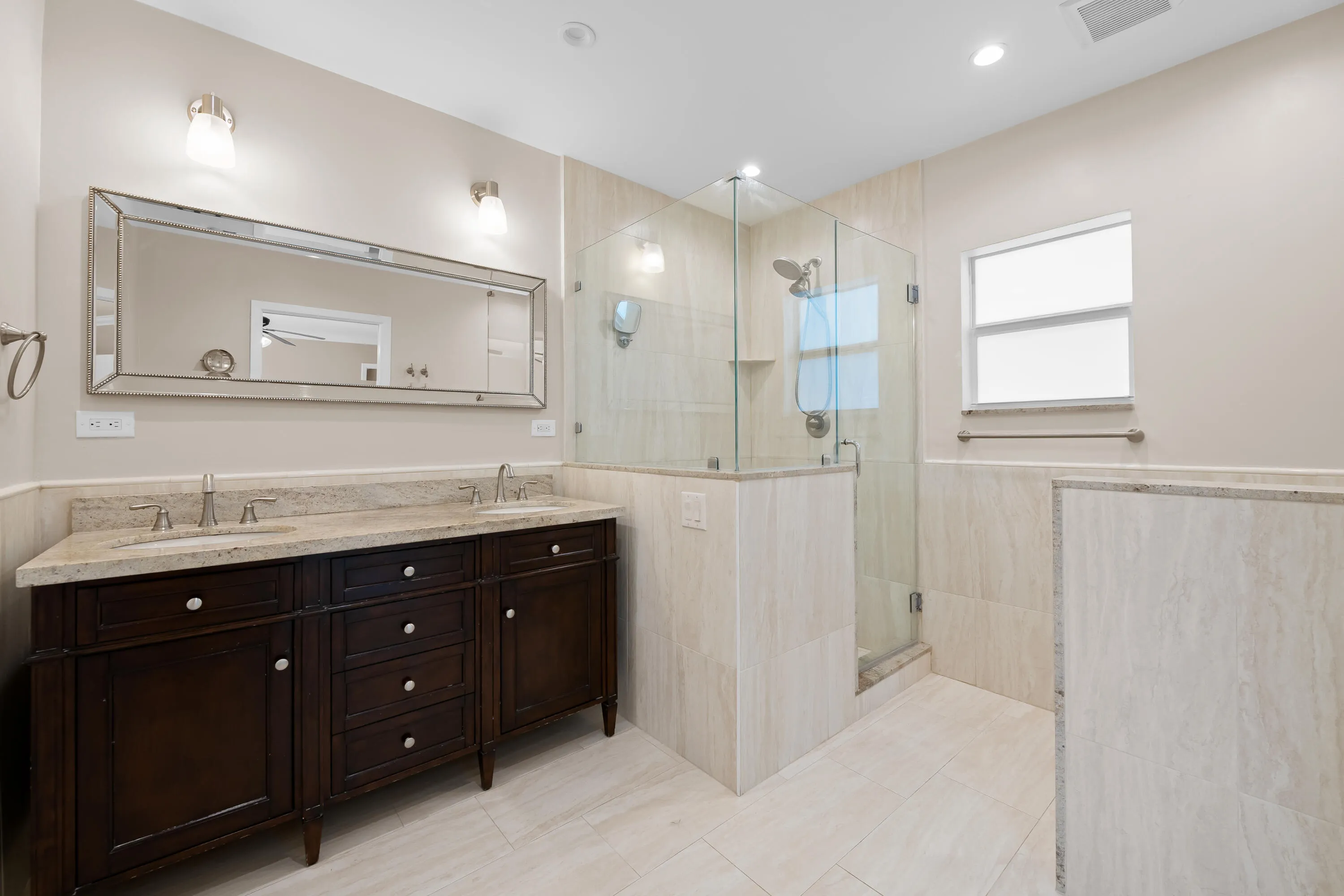 Dual-sink vanity with a light-toned countertop, a framed rectangular mirror, a glass-enclosed shower, and light-toned wall and floor tiling
