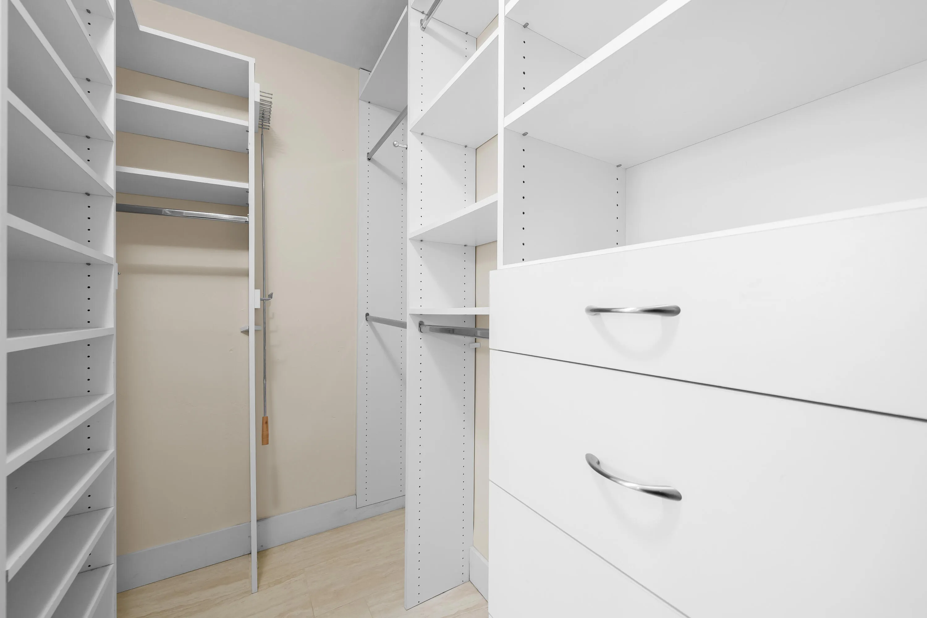 Custom closet system featuring white shelving units, integrated drawers with brushed chrome pulls, multiple hanging rods, and light-toned flooring