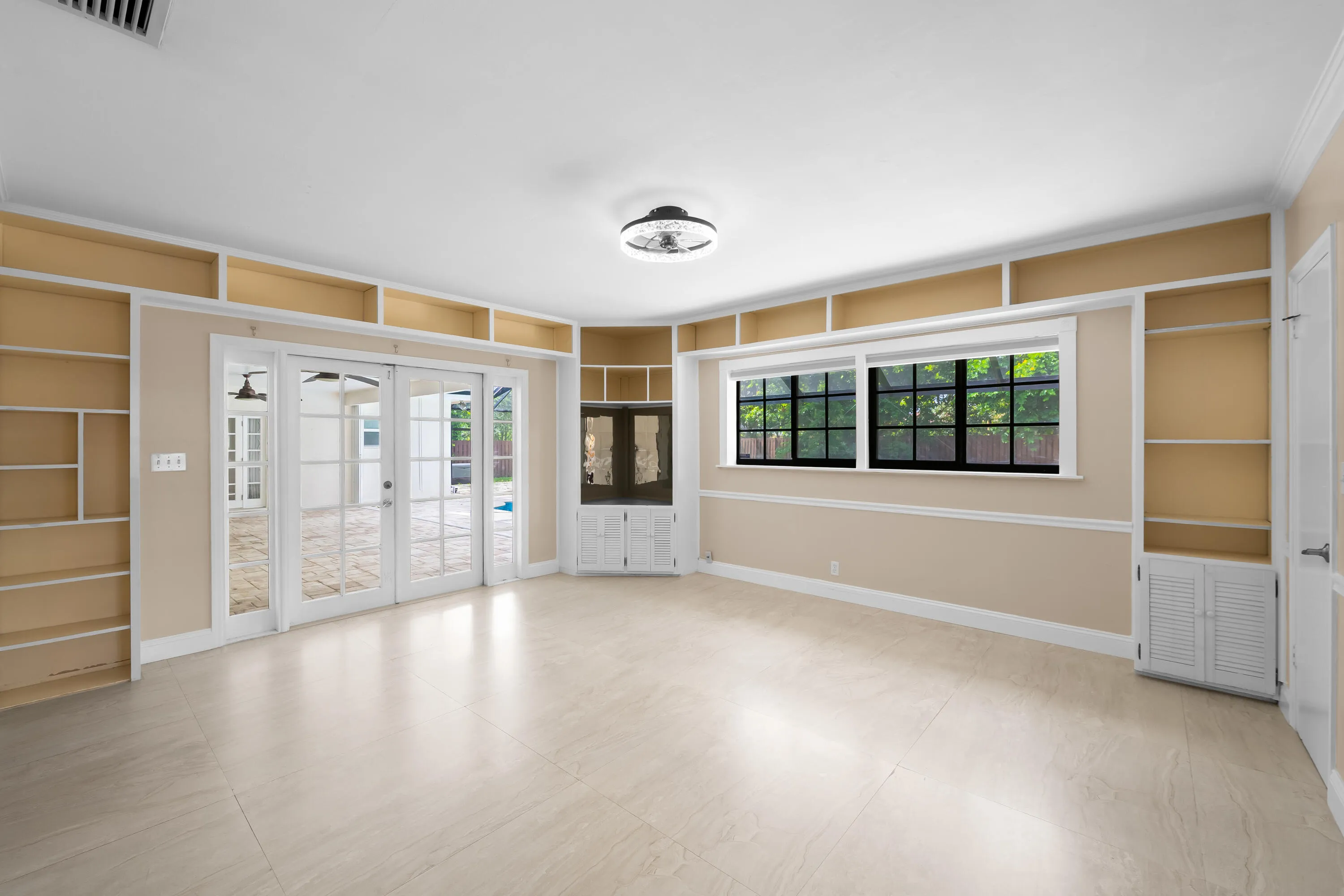 Spacious interior featuring light-toned tile flooring, extensive built-in shelving, a corner built-in cabinet, and large glass French doors opening to an outdoor area.