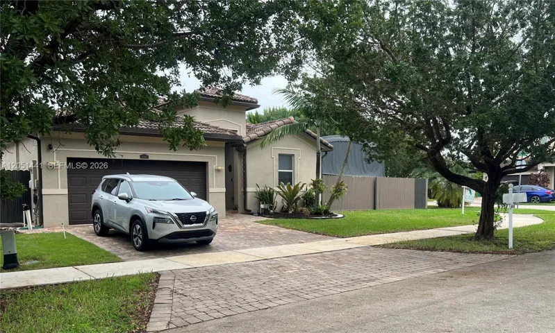 Stucco exterior featuring a paver driveway, two-car garage, tile roofing, and mature landscaping
