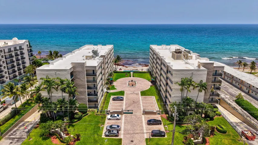 Oceanfront apartment buildings featuring private balconies, mature palm trees, manicured lawns, and a paver drive with central fountain
