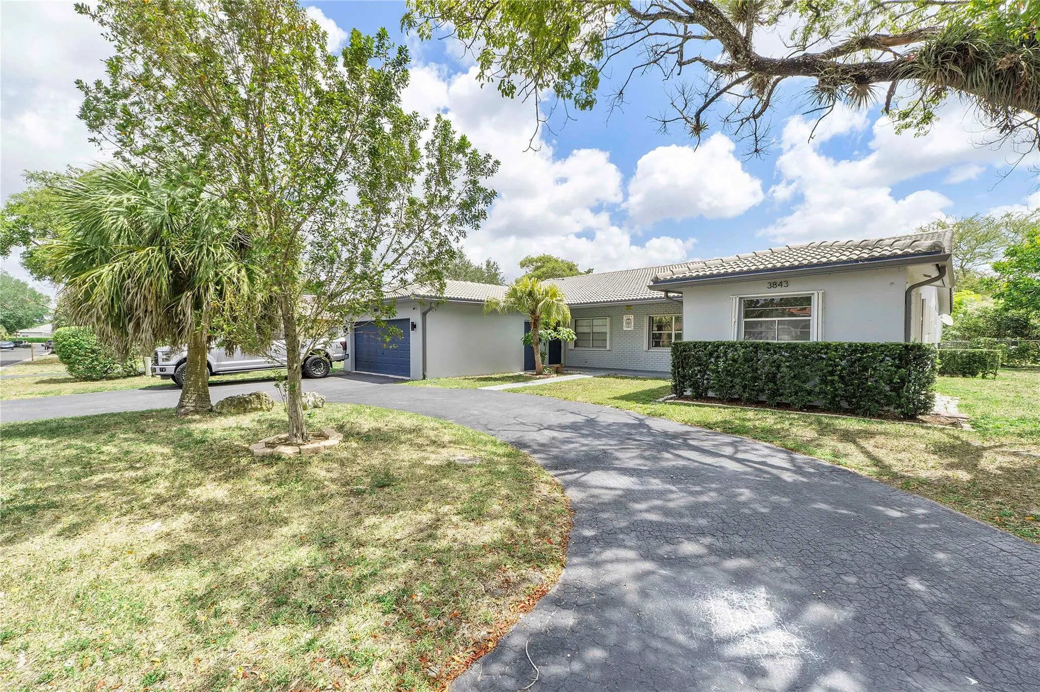 Contemporary stucco exterior with a tiled roof, mature tropical landscaping, a paved driveway, and an attached garage with a blue door