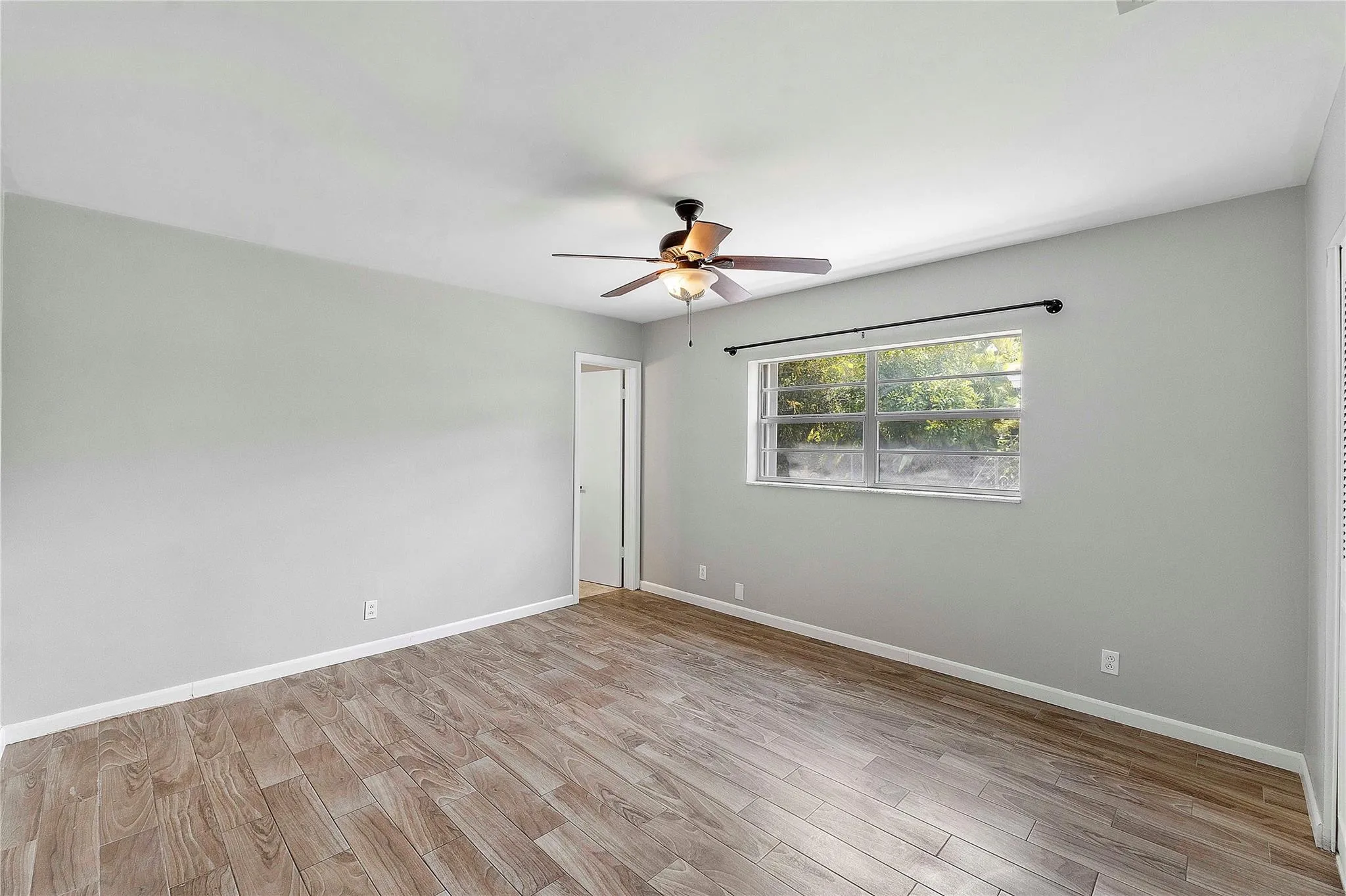 Spacious room featuring light wood-finish flooring, a ceiling fan, a large window, light gray wall paint, and white trim