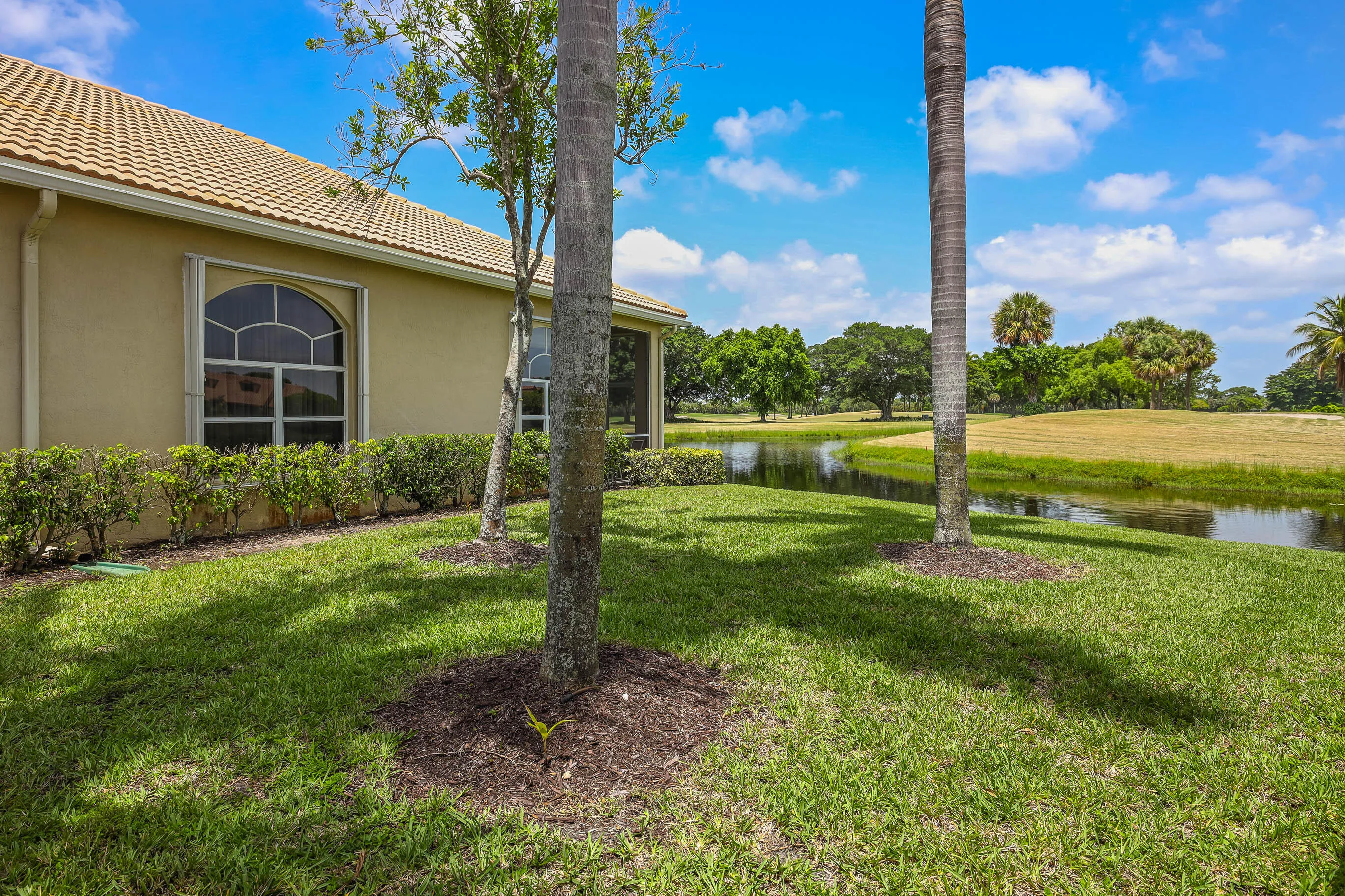 Manicured lawn with palm trees and a tranquil pond feature, bordered by a stucco exterior with an arched window and a tiled roof