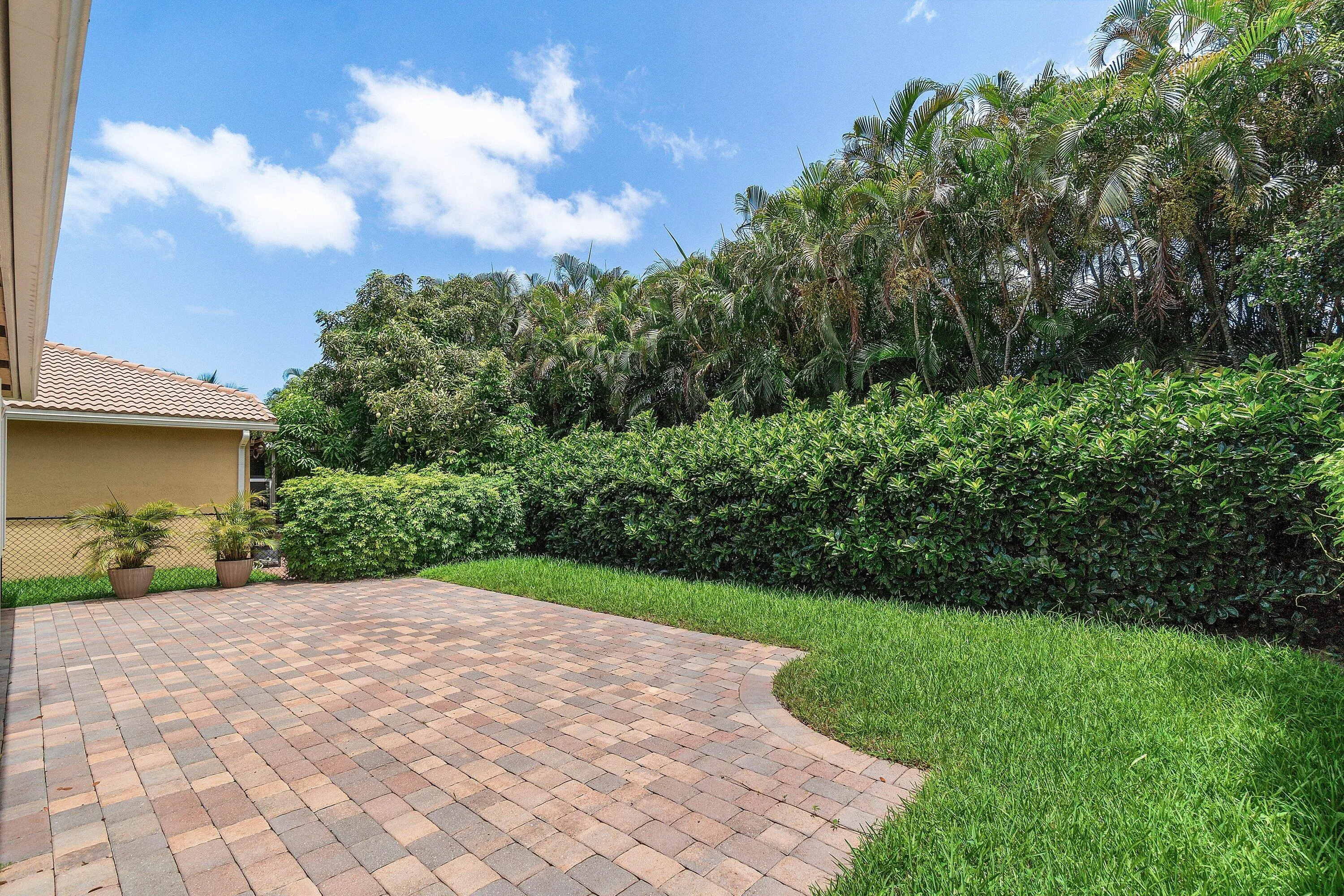 Spacious paver patio with a curved border, surrounded by lush green lawn and mature hedges. Property line extends beyond hedges. Room for a pool