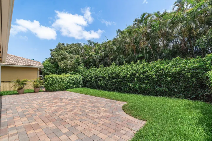 Spacious paver patio with a curved border, surrounded by lush green lawn and mature hedges. Property line extends beyond hedges. Room for a pool
