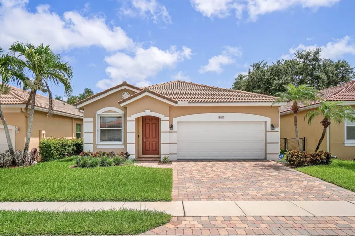 Single story home featuring a tile roof, a brick paver driveway, an arched entry door with decorative panels, a white garage door, and mature palm trees