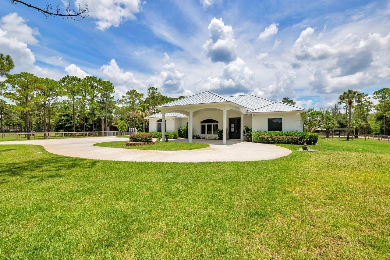 Spacious property featuring a white exterior residence with a covered entryway, a circular driveway, mature landscaping, and a surrounding privacy fence