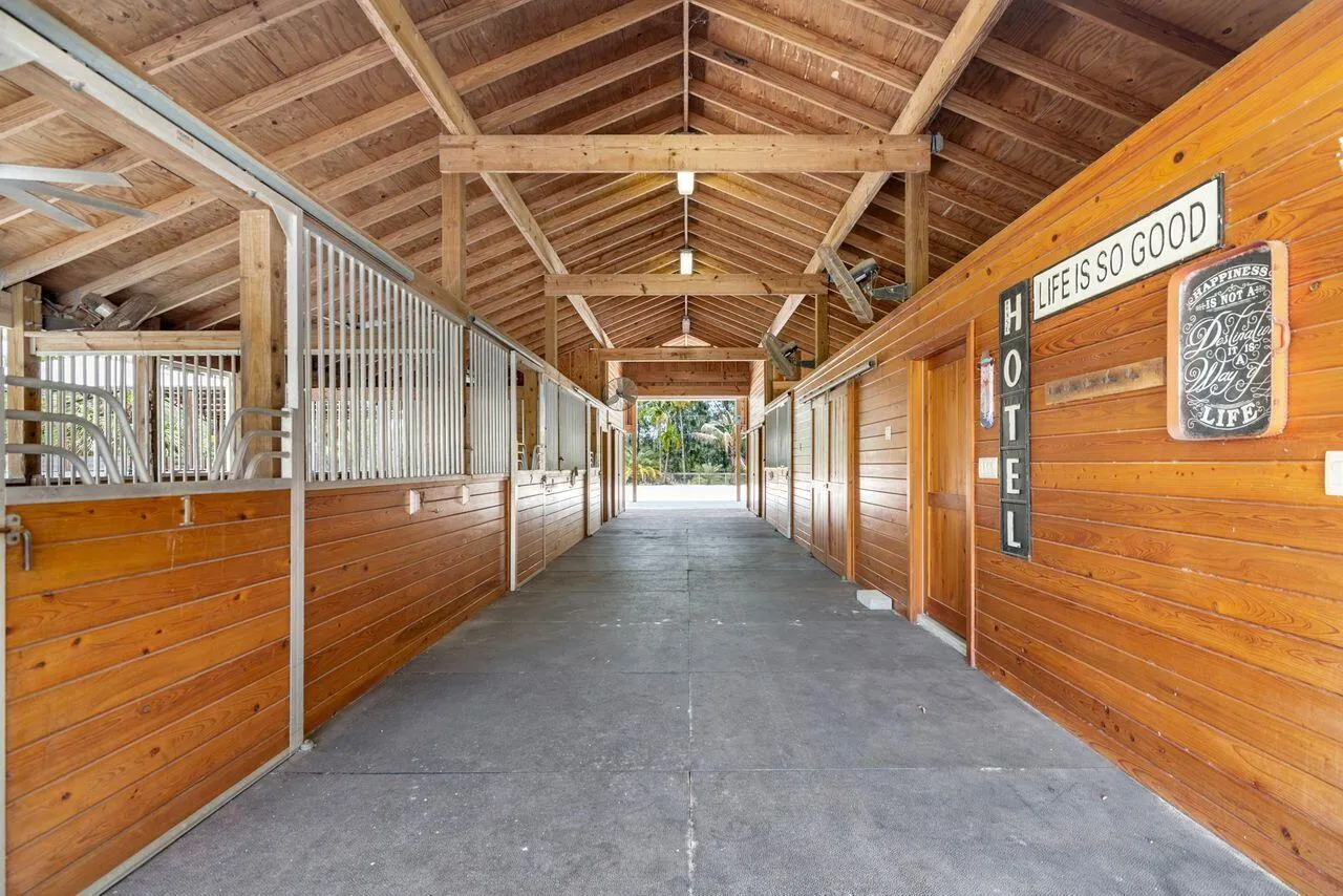 Spacious barn interior featuring exposed wood beams and ceiling, multiple stalls with custom stall doors, center isle, fans, fly system, 3 stalls have run outs.