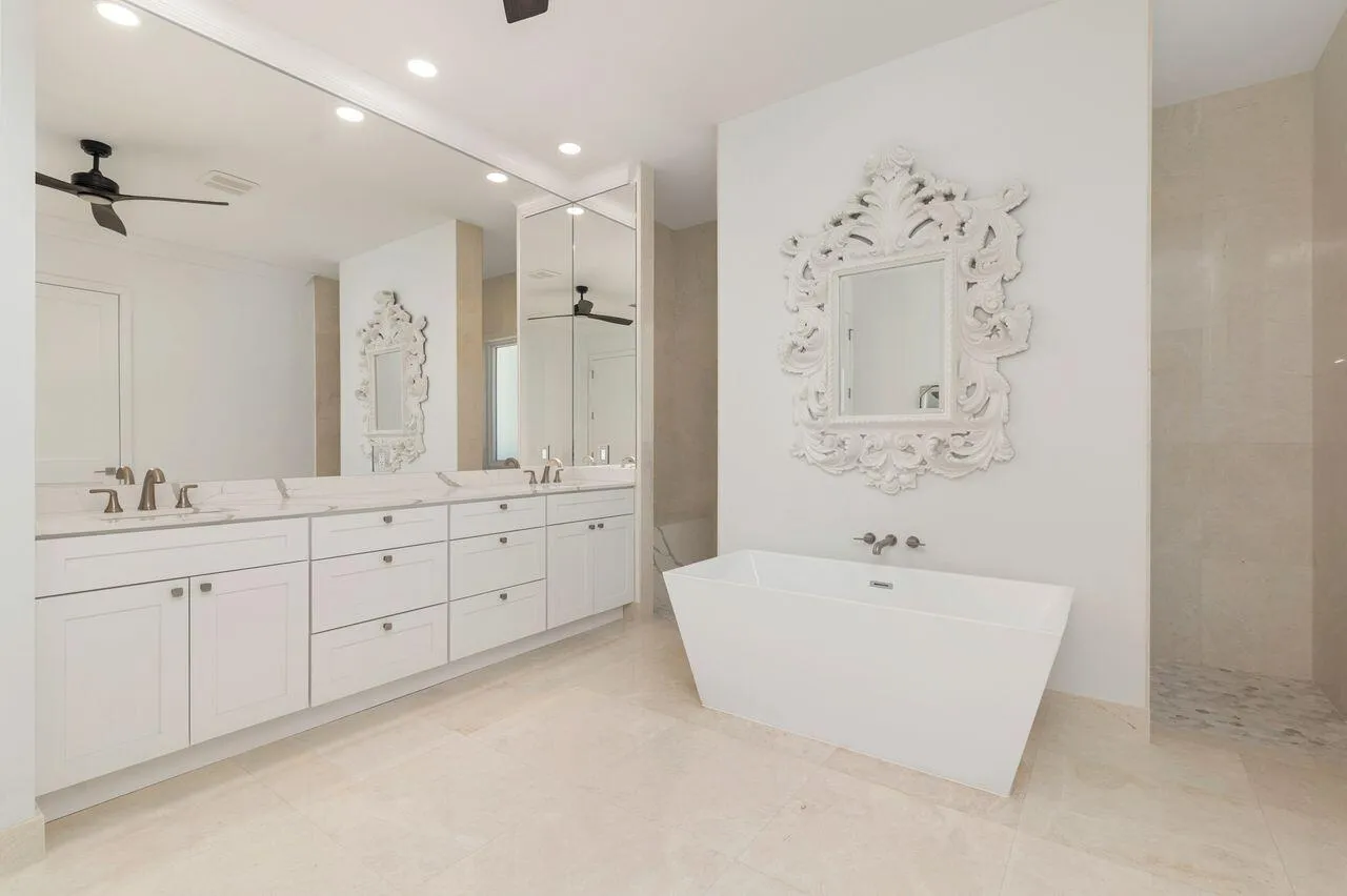 Spacious bathroom featuring a dual vanity with white cabinetry and light-toned countertops, a freestanding white soaking tub, and a glass-enclosed shower with pebbled flooring