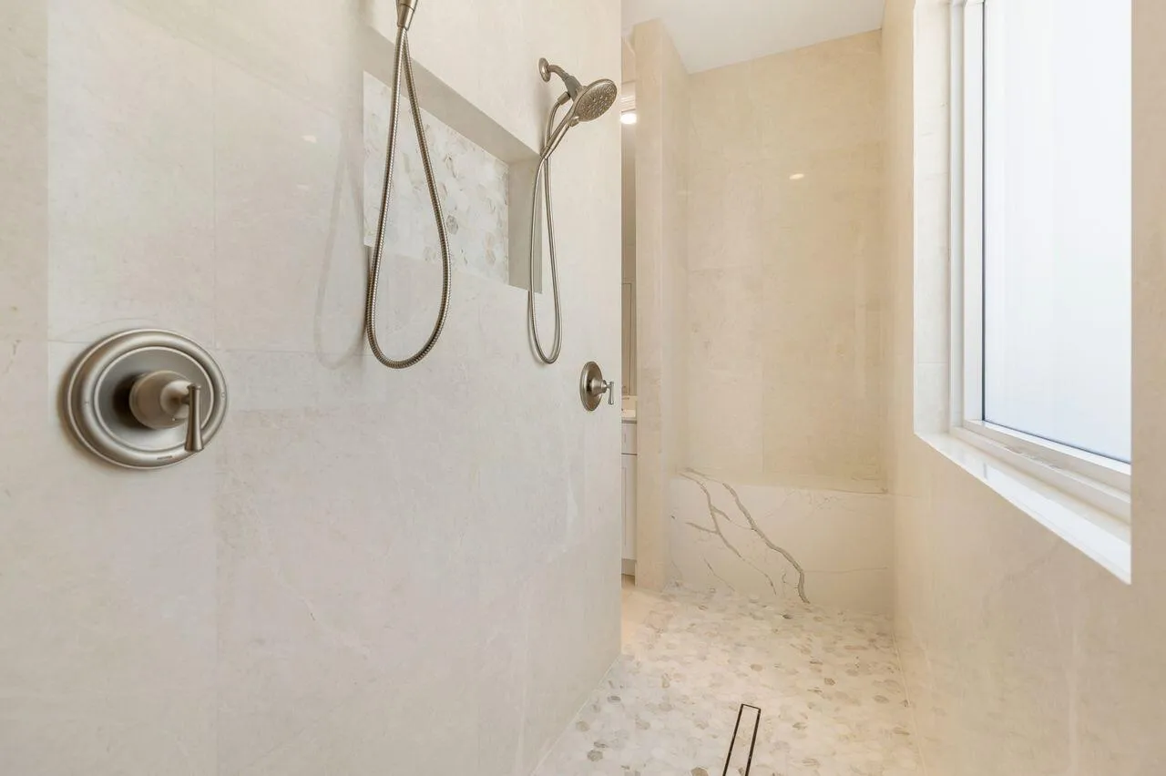 Spacious shower featuring light-toned tile walls, dual shower heads with brushed nickel fixtures, a built-in bench with veined stone, pebble-finish flooring, and a frosted​​‌​​​​‌​​‌‌​‌‌​​​‌‌​‌​‌​‌​​​‌​​ window
