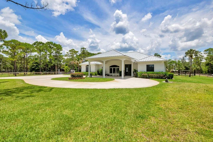 Spacious property featuring a white exterior residence with a covered entryway, a circular driveway, mature landscaping, and a surrounding privacy fence