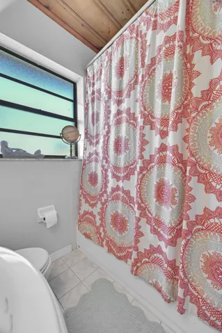 White tiled shower/tub combination with a window, wood-paneled ceiling, and light-toned floor tiles