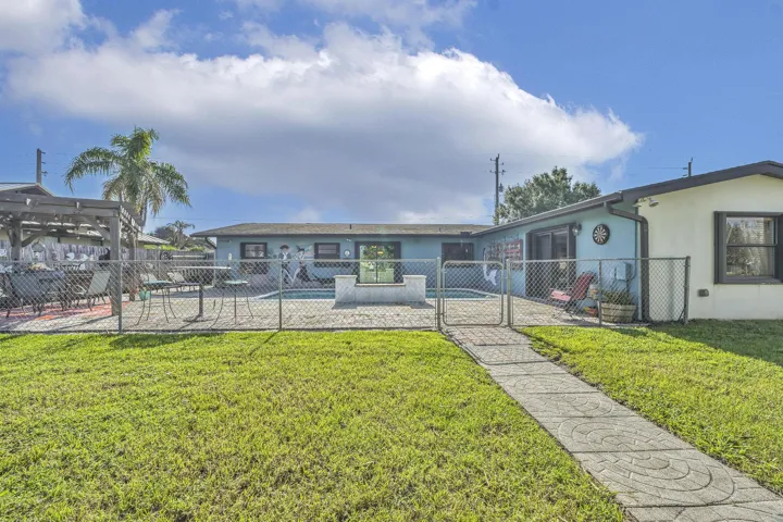 Expansive backyard featuring a manicured lawn, a concrete paver walkway, and a chain-link fence enclosing a concrete patio area with an in-ground pool