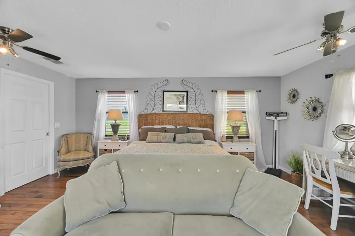 Spacious room featuring wood-finish flooring, two large windows with blinds, a white paneled door, and two ceiling fans with integrated lighting