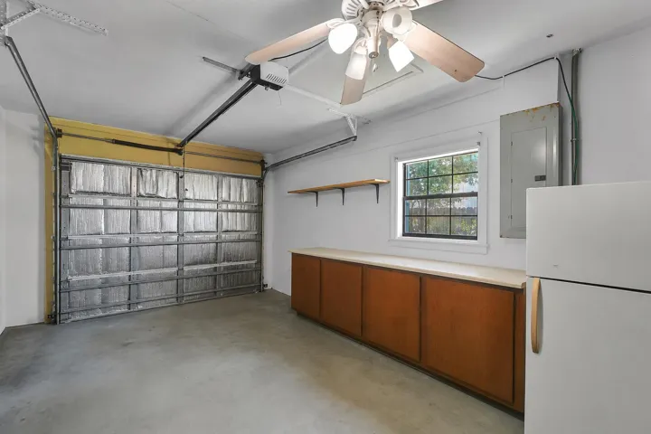 Garage featuring an insulated overhead door, concrete flooring, built-in wooden cabinetry with a light-toned countertop, a window, and a ceiling fan with integrated lighting