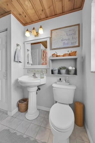 Pedestal sink with chrome faucet, light gray tile flooring, wood-paneled ceiling, and a built-in wall shelf